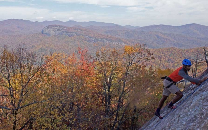 A person wearing safety gear is secured by ropes as they make their way up a rock incline. Behind them is a vast mountainous landscape covered in fall-colored trees. 